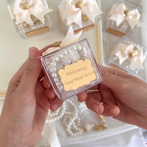 May include: Clear square favor boxes with white pearl beads and a gold label that reads "Welcoming Yusuf Hayat Baksh". Each box is tied with a cream-colored ribbon and a gold embellishment. Several boxes are visible.