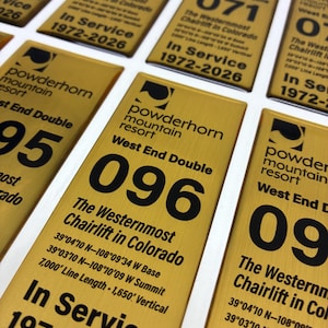 May include: A collection of rectangular gold-colored metal plaques featuring black text and logos for Powderhorn Mountain Resort. Each plaque displays a unique chairlift number, the text The Westernmost Chairlift in Colorado, coordinates, and In Service 1972-2026.