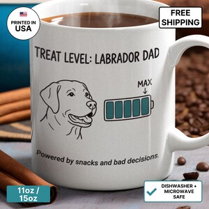 Puede incluir: Una taza de cerámica blanca con un dibujo de líneas negras de una cabeza de Labrador junto a un icono de batería lleno al máximo. El texto dice: TREAT LEVEL: LABRADOR DAD y Powered by snacks and bad decisions. La taza tiene un asa.