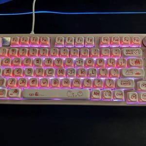Könnte beinhalten: Eine weiße Tastatur mit rosa Hintergrundbeleuchtung und benutzerdefinierten Tastenkappen mit Illustrationen von Comic-Tieren. Die Tastenkappen haben rote Beschriftungen und die Tastatur hat ein weiß-rosa Design.