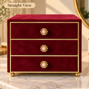 May include: A deep red velvet jewelry box featuring three horizontal drawers with gold-toned metal trim. Each drawer is equipped with a circular knob shaped like a flower with a central white pearl. The box stands on four small gold-toned feet.