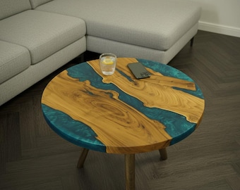 May include: A circular coffee table featuring a teal resin river design set within a wooden surface. The table has three wooden legs. A glass of water with lemon and a smartphone rest on the table. The table is approximately 76 cm in diameter.