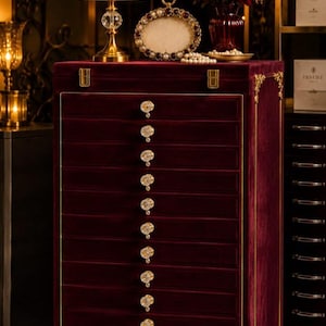 May include: A tall, rectangular jewellery chest upholstered in deep burgundy velvet with gold-toned metal corner accents. It features eleven horizontal drawers, each equipped with a round, faceted crystal knob. The top surface is flat.