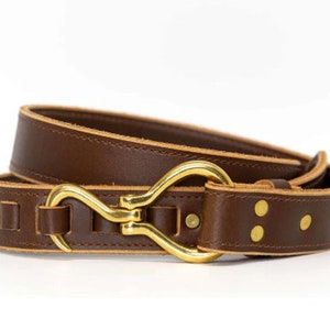 May include: A brown leather belt with a brass buckle in the shape of a hook. The belt has a stitched edge and two brass rivets.