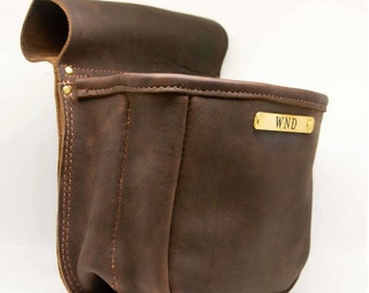 May include: A dark brown leather pouch with a flap closure. The pouch has a rectangular shape with a rounded bottom and a small gold-colored plate with the letters "WND". The leather appears to be distressed, with visible stitching.