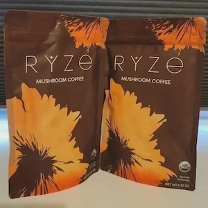 Pu&ograve; includere: Due buste marroni di caff&egrave; ai funghi Ryze con un grande disegno di un fiore arancione e giallo. Il testo riporta Ryze Mushroom Coffee, USDA Organic e Net Wt. 180g. La confezione include una chiusura a zip e un logo di certificazione bio.
