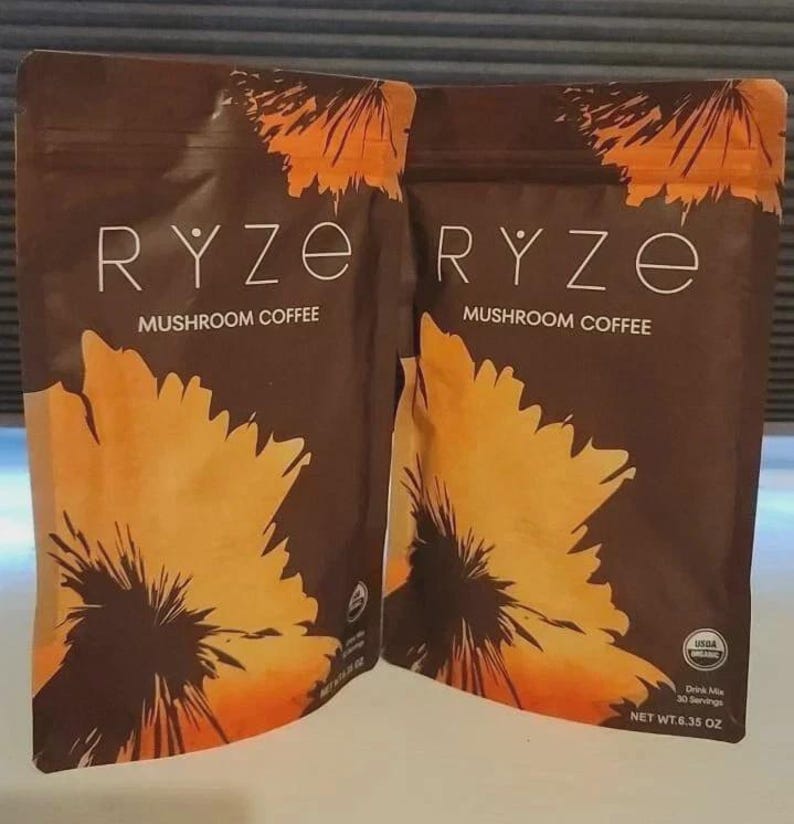 Pu&ograve; includere: Due buste marroni di caff&egrave; ai funghi Ryze con un grande disegno di un fiore arancione e giallo. Il testo riporta Ryze Mushroom Coffee, USDA Organic e Net Wt. 180g. La confezione include una chiusura a zip e un logo di certificazione bio.