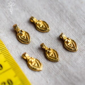 Könnte beinhalten: Fünf goldfarbene Metall-Anhänger in Form eines stilisierten Blattes mit einem erhabenen Design. Die Anhänger sind auf einer weißen Stoffoberfläche neben einem gelben Maßband angeordnet.