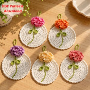 Könnte beinhalten: Fünf handgefertigte Häkeluntersetzer im floralen Design. Jeder Untersetzer hat eine weiße, kreisförmige Basis mit einer Blume in Rosa-, Orange-, Gelb- und Lilatönen. Die Untersetzer haben einen grünen Stiel und Blätter mit einer Schlaufe zum Aufhängen. Der Text "PDF Pattern download" ist sichtbar.