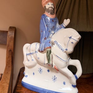 May include: A ceramic figurine depicting a figure on horseback. The rider wears a blue robe and a turban. The horse is white with blue accents. The base is white with blue floral designs and a blue border. The figurine is on a wooden surface.