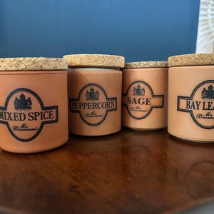Puede incluir: Cinco tarros de especias de cerámica marrón con tapas de corcho. Cada tarro tiene una etiqueta negra con una corona y el nombre de la especia: Romero, Especias Mixtas, Pimienta Negra, Salvia y Hojas de Laurel.