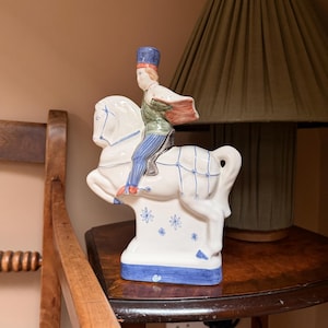 May include: A white ceramic figurine of a person riding a horse. The rider wears a blue hat, green jacket, and blue pants. The horse has blue accents and floral designs. The figurine is on a dark wooden surface.