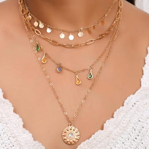 Puede incluir: Juego de collares dorados en capas. La capa superior presenta pequeños dijes redondos. La segunda capa tiene un diseño de eslabones de cadena. La tercera capa tiene dijes de lágrima coloridos. La capa inferior tiene un colgante de sol.