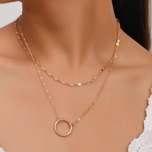 May include: A gold-colored, layered necklace with a delicate design. The top layer features small, rectangular links, while the lower layer has a simple circle pendant. The necklace is worn on a person, against a white top.