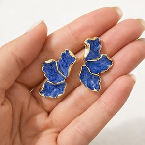 Könnte beinhalten: Ein Paar blau-goldener Ohrringe. Jeder Ohrring hat ein blaues, blütenblattförmiges Design mit goldenem Rand. Die Ohrringe sind vor einem neutralen Hintergrund platziert.