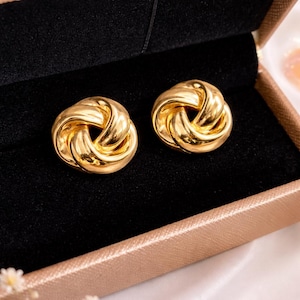May include: A pair of gold-colored knot-shaped stud earrings. The earrings are displayed in a black velvet jewelry box with a gold-colored trim. The earrings have a polished, reflective surface.