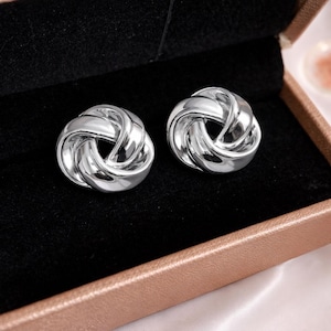 Peut inclure: Une paire de boucles d'oreilles argentées. Chaque boucle d'oreille présente un design noué, avec des surfaces lisses et réfléchissantes. Les boucles d'oreilles sont présentées dans un écrin à bijoux doublé de velours noir avec un extérieur beige.