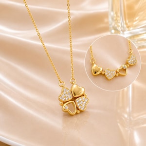 Puede incluir: Un collar de tono dorado con un colgante en forma de trébol de cuatro hojas compuesto por cuatro corazones. Dos corazones son de metal dorado liso y dos corazones están incrustados con pequeñas piedras transparentes. La cadena es fina.