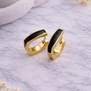 May include: A pair of gold-colored hoop earrings with a rectangular shape. Each earring features a thick, black enamel stripe across the top. The earrings are displayed on a marble surface, with a white cloth and dried flowers in the background.