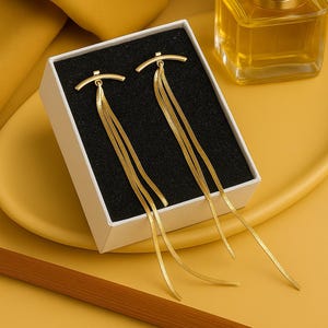 May include: Gold-toned earrings with a linear design, presented in a white box with a black interior. The earrings feature a curved top and multiple dangling chains. A gold-colored perfume bottle is in the background.