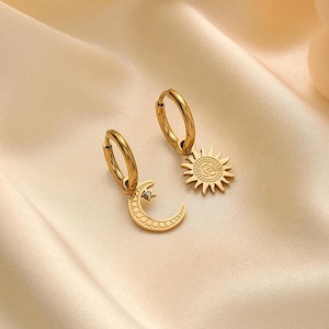 May include: A pair of gold-colored hoop earrings. One earring features a crescent moon charm with a small crystal, and the other has a sun charm. The earrings are displayed on a cream-colored, satin-like fabric.