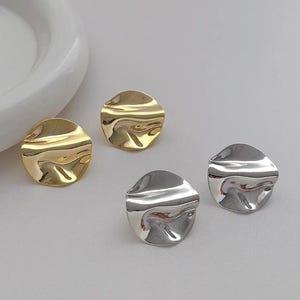 May include: Four round earrings, two in gold and two in silver. The earrings have a textured, rippled surface, reflecting light. The earrings are a simple, yet elegant design.