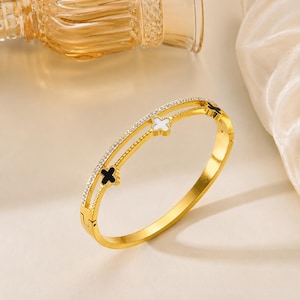 Könnte beinhalten: Ein goldfarbenes Klapparmband mit doppeltem Banddesign. Ein Band ist mit kleinen, klaren Steinen besetzt, während das andere abwechselnd schwarze und weiße vierblättrige Kleemotive mit glatten, goldfarbenen Rändern aufweist.