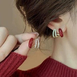 May include: Gold-coloured double hoop earrings. One hoop is encrusted with small, clear crystals, while the other is smooth gold. The earrings have a curved design and are being held up by a person with red nail polish.