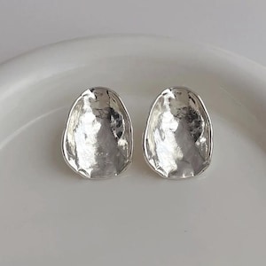 Puede incluir: Un par de pendientes de botón plateados y texturizados. Cada pendiente tiene una forma irregular y orgánica con una superficie reflectante. Se muestran sobre una superficie blanca, resaltando su brillo metálico.