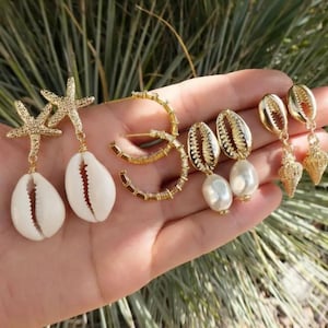 Puede incluir: Una colección de pendientes dorados con diseños de conchas marinas y estrellas de mar. Los pendientes incluyen estilos de aro, conchas colgantes con perlas y estrellas de mar con colgantes de concha blanca. Las joyas se exhiben sobre un fondo verde borroso.