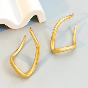 Puede incluir: Un par de pendientes de aro dorados y abstractos. Los pendientes tienen un diseño angular único con una parte superior curva y una inferior recta. Los pendientes se muestran sobre una superficie azul claro.