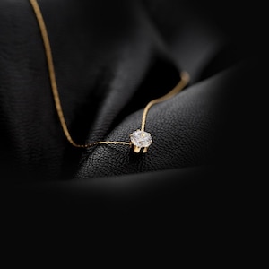 Puede incluir: Un delicado collar de oro con un único colgante de diamante brillante. El collar se muestra sobre un fondo oscuro y texturizado, resaltando el brillo de la gema. La cadena es fina y elegante.
