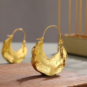 Puede incluir: Un par de pendientes de aro dorados con un diseño texturizado y orgánico. Los pendientes tienen forma de media luna con apariencia de metal martillado. Se exhiben sobre una superficie de madera.