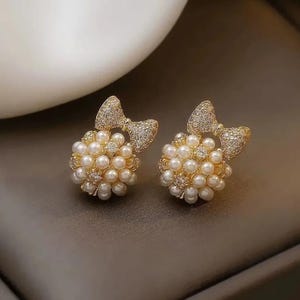 Puede incluir: Un par de pendientes de botón dorados. Cada pendiente presenta un grupo de pequeñas perlas redondas de color crema. Un adorno en forma de lazo, incrustado con cristales transparentes, se encuentra encima del grupo de perlas. Los pendientes están sobre un fondo neutro.