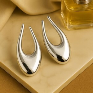 May include: A pair of silver-coloured, teardrop-shaped earrings with a polished finish. The earrings are displayed on a beige surface, with a perfume bottle in the background. The earrings have a modern, minimalist design.