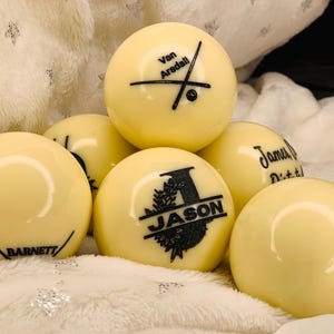 May include: Six white billiard balls with black personalized text. The balls are arranged in a pyramid shape. The text on the balls includes "Van Arsdell", "Jason", "James Dittrich", and "Barnett".