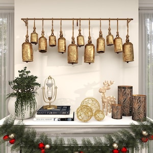 May include: A horizontal metal rod mounted on a wall with twelve hanging gold-toned metal bells of varying sizes. Below, a white mantel displays a potted green plant, a gold hourglass, two stacked books, three gold wire spheres, a light-up deer, and three cylindrical candle holders.