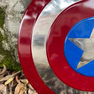 Op de afbeelding: Een rood, wit en blauw Captain America-schild met een zilveren metalen rand. Het schild ligt op een bed van bruine bladeren.