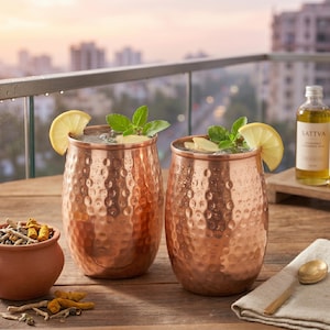 Puede incluir: Dos vasos de cobre martillado llenos de hielo, decorados con hojas de menta verde fresca y rodajas de limón amarillo. Un pequeño cuenco de terracota con trozos de cúrcuma y una botella de vidrio con la etiqueta SATTVA sobre una superficie de madera.