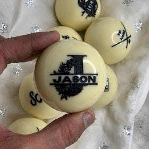 May include: Close-up of a white billiard ball with a black design that says "JASON" with a decorative floral border and the letter "I" in the center.