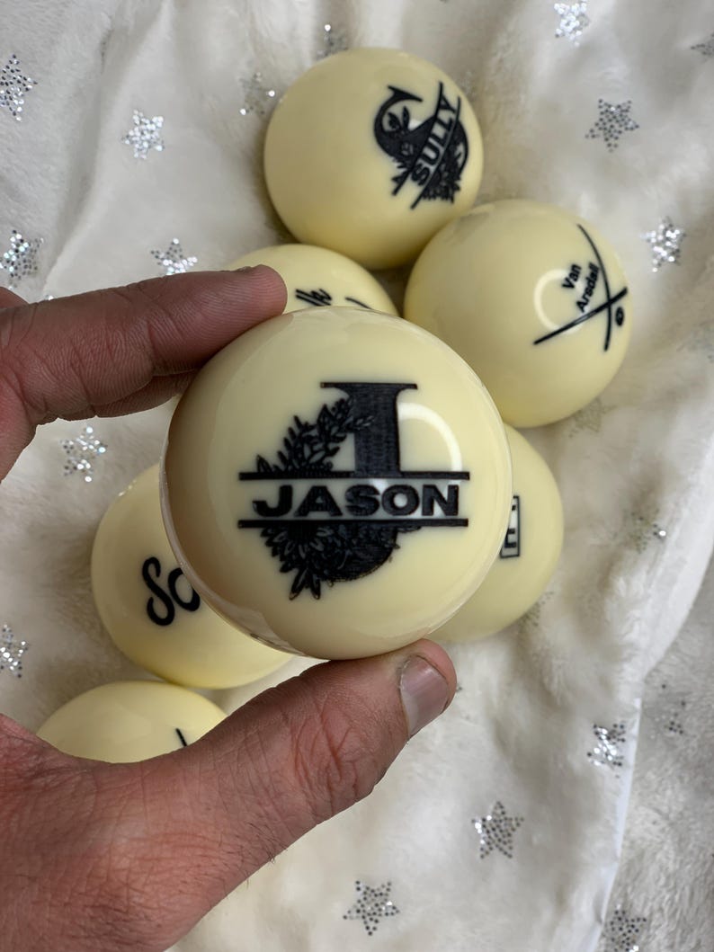 May include: Close-up of a white billiard ball with a black design that says "JASON" with a decorative floral border and the letter "I" in the center.