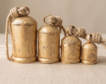 Può includere: Un set di quattro campane cilindriche graduate con una finitura color oro anticato. Ogni campana presenta una parte superiore arrotondata con un anello in metallo fissato a una spessa corda di juta color naturale, creando un aspetto rustico.