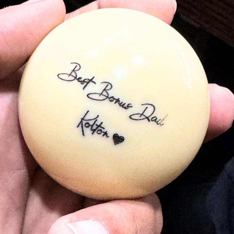 May include: A white billiard ball with black lettering that reads "Best Bonus Dad" and "Kotton" with a small heart symbol.