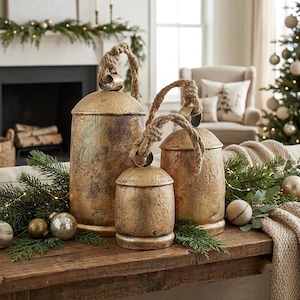 May include: Three rustic, gold-toned metal bells of varying sizes, each featuring a weathered, textured finish and a thick, natural jute rope handle attached to the top loop. The bells are arranged together, showcasing their cylindrical shapes.