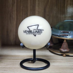 May include: A cream-colored billiard ball with the text "VENBILLIARD CORPORATION" and a logo. The ball is displayed on a black metal stand with a circular base. The background includes a wooden surface and a decorative metal object.