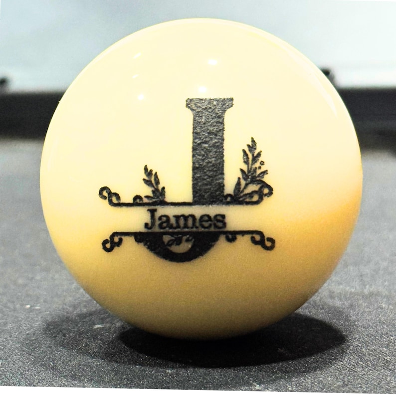 May include: A white billiard ball with a black monogram design. The design features the letter "J" with a banner that reads "James".
