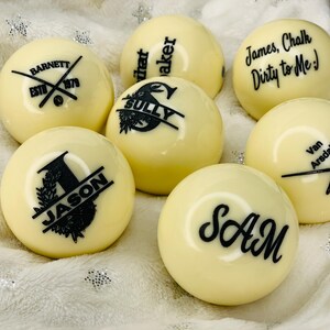 May include: Six white billiard balls with black personalized text. The text on the balls includes names, a company logo, and a funny saying.