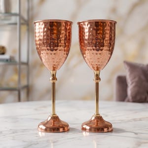 May include: Two matching goblets featuring hammered copper bowls, slender gold-toned stems with a central spherical accent, and circular copper bases. The metallic surfaces reflect light, highlighting the textured finish of the bowls.