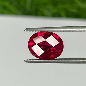May include: A faceted, oval-shaped, deep red gemstone held by silver tweezers. The gem has a brilliant cut, reflecting light with a starburst pattern. The background is a blurred green and white.