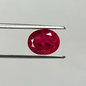 May include: A vibrant, oval-cut ruby held by silver tweezers. The gemstone displays a deep red hue, with visible facets catching the light. The background is a soft white, with a blurred green area at the top.